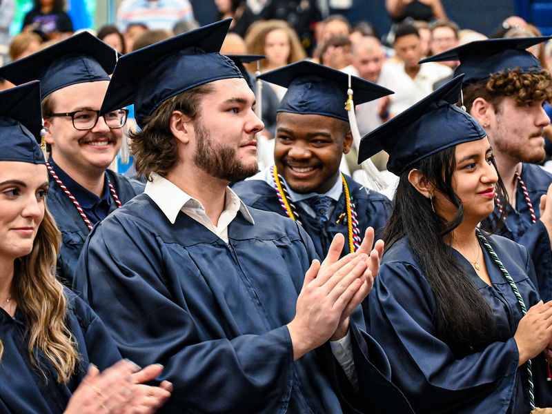 Close-up shot capturing the joyous moment of graduates, adorned in their blue caps and gowns, clapping and smiling during their graduation ceremony, radiating pride and accomplishment.