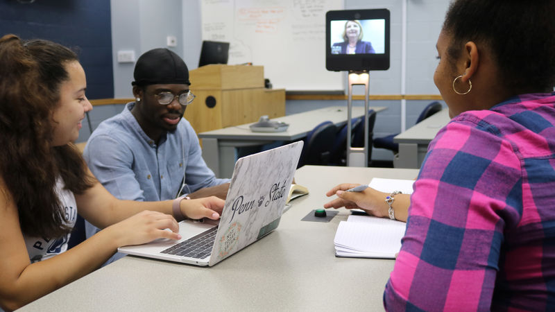 Three students learning from a professor on a lcd monitor attached to a robot. 