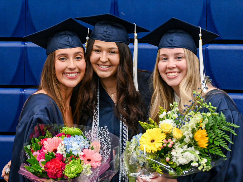 3 female graduates smiling for a photo while holding flowers at Penn State Greater Allegheny's Spring 2024 Commencement Ceremony