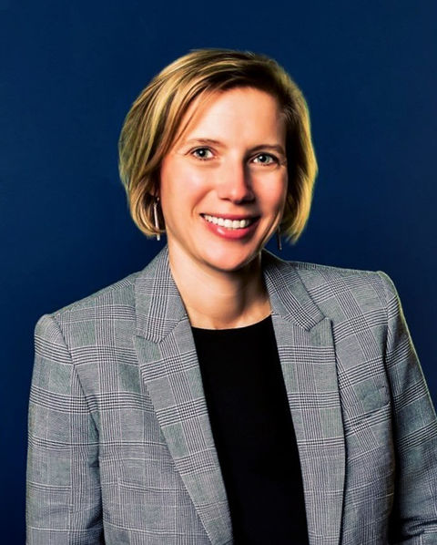 Photo of woman smiling wearing a grey blazer against a blue background.