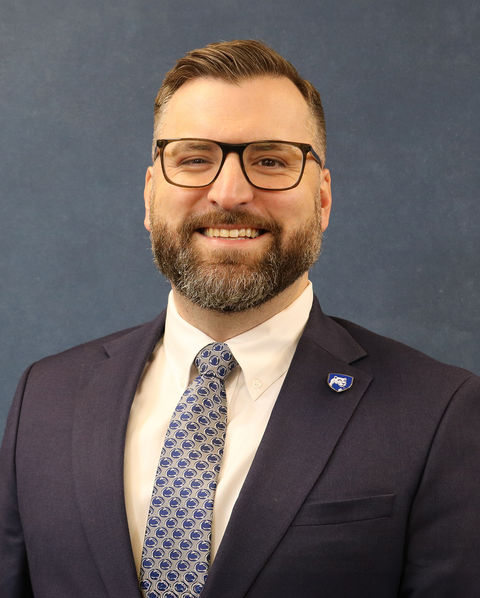 Man smiling wearing a suit with a Penn State shield pin, sat in front of a blue background