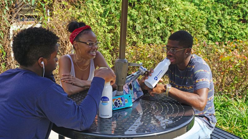 Three students sitting around a circular table outside. One student is holding their white water bottle displaying Greater Allegheny Logo 