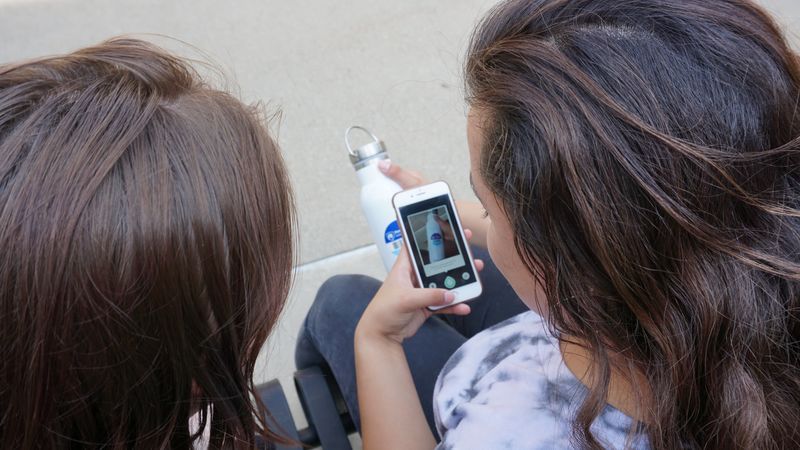 Two students sitting. One student is holding a phone while scanning a barcode that is on her water bottle