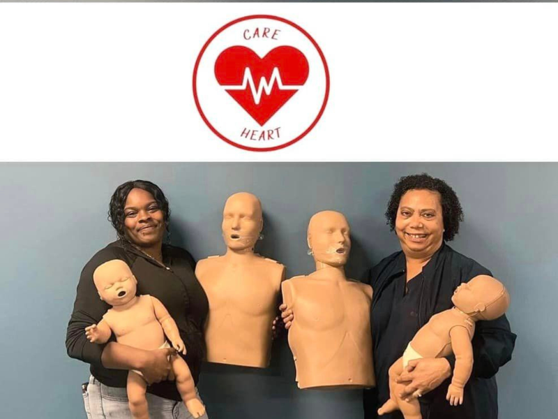 Two individuals stand in front of Care Heart CPR logo holding CPR dummies