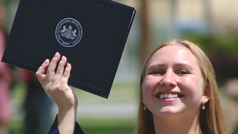 Penn State graduate holds up diploma and smiles while outdoors