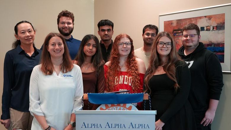 Smiling students and faculty members stand together behind an Alpha Alpha Alpha (Tri-Alpha) Honor Society podium