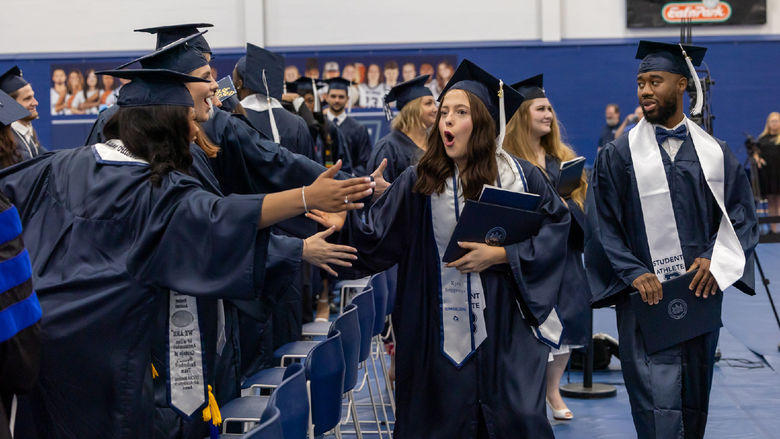 A graduate greets 2 friends as she exits after commencement. All are dressed in caps and gowns.