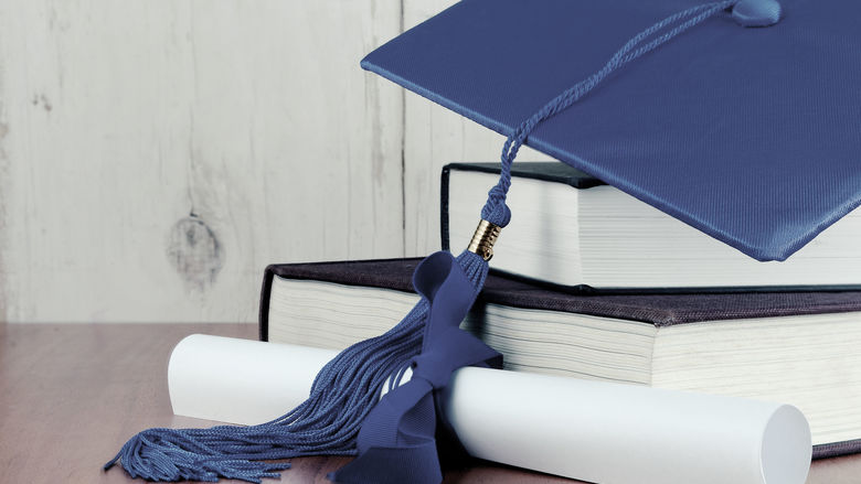 A graduation cap on two books with a diploma tied with blue ribbon.
