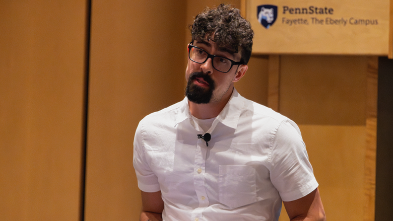Allain Daigle sits with a laptop in his lap, speaking during a faculty development session at Penn State Fayette. A Penn State logo and campus mark are visible in the background.