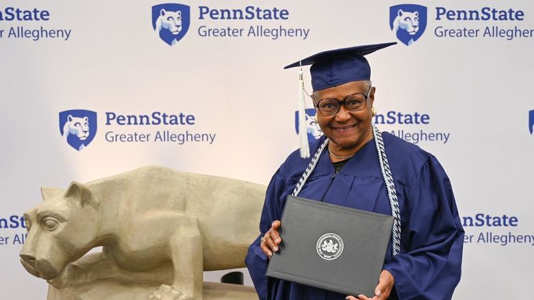 Diane Banks, wearing a graduation cap and gown, smiles while holding her diploma in front of the Penn State Greater Allegheny backdrop and Nittany Lion statue.