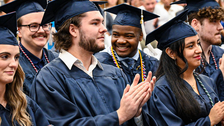 Close-up shot capturing the joyous moment of graduates, adorned in their blue caps and gowns, clapping and smiling during their graduation ceremony, radiating pride and accomplishment.