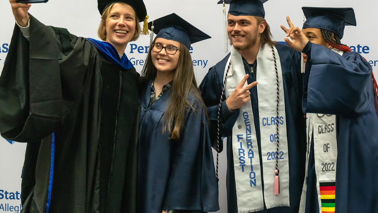 Megan L. Nagel in academic regalia takes a selfie with three graduates in caps and gowns, all smiling and posing in front of a Penn State Greater Allegheny step-and-repeat backdrop.