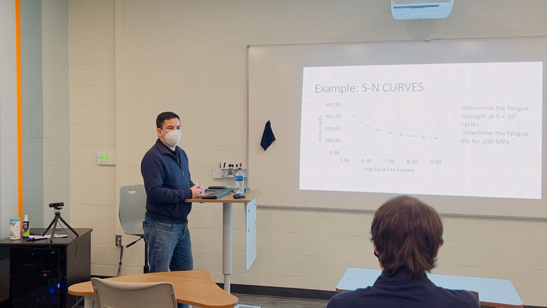 Man standing at front of room behind a podium. A projector projects a chart on a white board. In the forefront of the photo there is a student looking at the man.