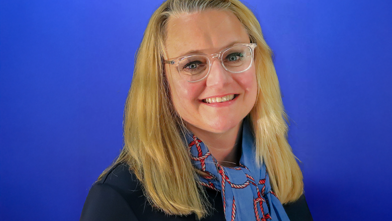 Jennifer Breese, smiling, wearing glasses and a blue scarf with a red rope pattern, standing in front of a blue background.