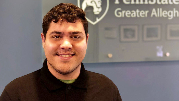 Student Derrick Ferrer smiling in front of Penn State Greater Allegheny logo
