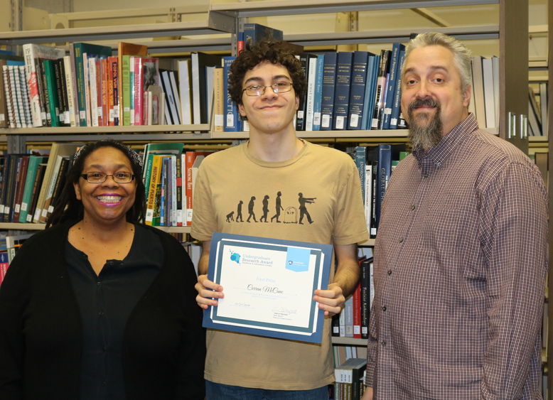 Three people standing in front of a bookshelf smiling. The person in the middle is holding an award. 