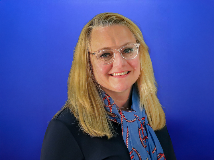 Jennifer Breese, smiling, wearing glasses and a blue scarf with a red rope pattern, standing in front of a blue background.